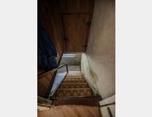 Blick in ein schmales, alt wirkendes Treppenhaus mit hölzerner Decke und abgenutzten Putzwänden. Eine steile Treppe mit gemustertem Belag und Holzgeländer führt hinunter zu einem Kellerbereich mit freiliegendem Mauerwerk und kleinem Fenster.