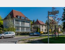 Wohngebiet mit einer Kreuzung, an der zwei Straßenschilder die Namen „Ravenweg“ und „Oertgenweg“ anzeigen. Im Vordergrund ein geparkter silberner Minivan, daneben ein gelber Briefkasten, und im Hintergrund Häuser mit roten Giebeldächern sowie blühende Bäume.