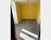 Ein leerer Raum mit gelben Wänden und einem Holzfußboden. An der Decke hängt ein einfacher, goldfarbener Kronleuchter mit mehreren Armen, und natürliches Licht fällt durch ein Fenster auf den Boden.