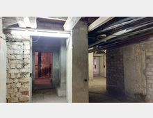 Ein unsanierter Kellerraum mit rohen Beton- und Backsteinwänden, sichtbaren Balken und Rohrleitungen an der Decke. Im Hintergrund sind weitere Räume und ein rötlich beleuchteter Bereich zu sehen.