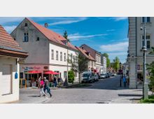 Straßenszene in einer Kleinstadt mit historischen Gebäuden, gepflasterter Straße und kleinen Geschäften. Vor einem Geschäft mit roten Sonnenschirmen laufen Fußgänger, während Autos entlang der Straße parken.