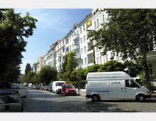 Straßenansicht einer Wohngegend mit mehrstöckigen weißen Altbauten und Balkonen, flankiert von Bäumen. Auf der gepflasterten Straße parken mehrere Autos und Lieferwagen.