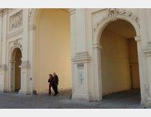 Sandfarbener, klassizistischer Torbogen mit dekorativen Reliefs, flankiert von massiven Säulen. Im Vordergrund gehen zwei Personen auf einem gepflasterten Weg entlang.