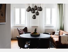 Helles Wohnzimmer mit einem runden Esstisch aus dunklem Holz, umgeben von schwarzen Stühlen. Im Hintergrund befinden sich zwei große, weiß umrahmte Fenster mit halbtransparenten Vorhängen, eine braune Sofaecke und ein Ledersessel; eine moderne Lampe mit glänzenden Kugellichtern hängt von der Decke.