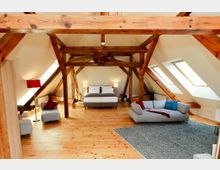 Helles Dachgeschosszimmer mit freiliegenden Holzgebälken, einem großen Doppelbett in der Mitte, einer grauen Couch auf einem Teppich und schrägen Wänden mit Dachfenstern. Der Raum ist mit Holzboden und moderner Beleuchtung ausgestattet.