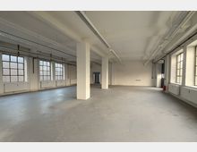 Großer, leerer Industrie-Loft-Raum mit Betonboden, weißen Stützsäulen und hohen Decken mit sichtbaren Kabeltrassen und langen Leuchten. Entlang der Wände befinden sich große, rasterförmige Fenster und Heizkörper; im Hintergrund ist eine Türöffnung zu sehen.