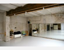 Ein heller Tanz- oder Übungsraum mit weißem Boden, freiliegenden Ziegelwänden und rohen Deckenbalken. Eine große Spiegelwand dominiert die rechte Seite, daneben sind gelagerte Matten, Decken und Stühle sichtbar.