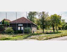 Ein kleines, eingeschossiges Gebäude mit brauner Holzfassade und rotem Ziegeldach, umgeben von einem Zaun und Bäumen, steht in einer ländlichen Umgebung. Im Vordergrund ist ein ungepflasterter Weg und im Hintergrund sind Wiesen zu sehen.