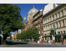 Straßenszene mit historischem Gebäude, das die goldene Kuppel der Neuen Synagoge zeigt, flankiert von Bäumen und alten Wohn- und Geschäftshäusern; im Vordergrund Gehweg, Fahrradständer und ein Straßenschild.
