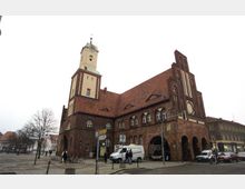 Backsteingebäude mit historischem Rathauscharakter, bestehend aus einem markanten Uhrturm und roten Ziegeldächern. Im Vordergrund befindet sich ein kleiner Platz mit Fußgängern, Bäumen und geparkten Fahrzeugen.