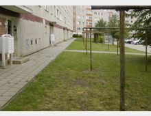 Eine Außenaufnahme eines mehrstöckigen Wohnblocks mit grauer Fassade. Rechts davon liegt eine Rasenfläche mit Wäscheleinen und im Hintergrund sind weitere Gebäude sowie geparkte Autos zu sehen.