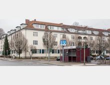 Mehrstöckiges Wohngebäude mit heller Fassade, roten Ziegeldächern und blauen Fensterläden, im Vordergrund eine geschlossene Imbissbude mit dem Schild "Berliner Imbiss" und ein U-Bahn-Schild für die Linie U6/U7. Die Szene zeigt eine städtische Straßenecke mit kahlen Bäumen, Verkehrsampeln und parkenden Autos.