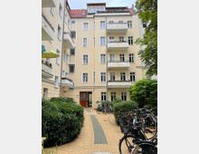 Innenhof eines mehrstöckigen Wohnhauses mit hellgelber Fassade, weißen Fensterrahmen und mehreren Balkonen sowie rotem Ziegeldach. Ein gepflasterter Weg führt zum Hauseingang, flankiert von Grünflächen und Fahrradständern mit abgestellten Fahrrädern.