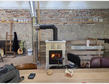 Ein rustikaler Innenraum mit einer freiliegenden Ziegelwand und einer Holzofen-Heizung, deren Schornstein nach oben führt. Im Vordergrund ein Holztisch mit Gegenständen wie einem Brot und einer Snackschale, im Hintergrund Malutensilien, eine Gitarre und verschiedene Möbelstücke.