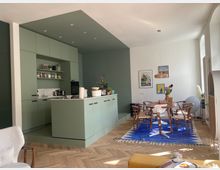 Helle Wohnung mit offener Küche und Essbereich. Die Küche hat grüne Einbauschränke und eine freistehende Kücheninsel, der Essbereich ist mit einem runden Holztisch, Stühlen und einem blauen Teppich eingerichtet.