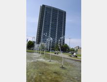 Hohes Gebäude der Technischen Universität mit moderner Glas- und Stahlfassade, im Vordergrund ein flaches Wasserbecken mit sprudelnden Fontänen. Umgeben von einer Grünfläche mit Bänken, auf denen Menschen sitzen, und Bäumen im Hintergrund.