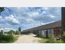 Langer, einstöckiger Gebäudekomplex mit einem grauen Ziegeldach auf einem ländlichen Gelände, umgeben von grüner Vegetation und einem unbefestigten Weg. Rechts sind mehrere Frachtcontainer aufgereiht, und links führt eine Einfahrt zu einer umzäunten Fläche.