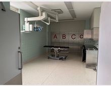 Ein steriler Operationssaal mit einer OP-Liege in der Mitte, einer Deckenlampe mit Beleuchtungseinheit und hinteren Wandschränken mit den Buchstaben „A“, „B“ und „C“. Rechts befindet sich eine Edelstahl-Arbeitsfläche mit Spüle.