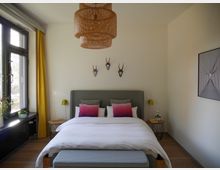 Schlafzimmer mit einem grauen Bett, farbigen Kissen und einer weißen Bettdecke. An der Wand hängen kleine Deko-Geweihe, rechts ein gerahmtes Bild, links gelbe Vorhänge vor einem großen Fenster; eine geflochtene Deckenlampe und zwei Nachttische mit Leuchten ergänzen das Design.