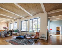 Helles Wohnzimmer mit großen bodentiefen Fenstern, gewölbter Ziegeldecke und Holzboden. Der Raum ist mit modernen Möbeln, einem Bücherregal, einem Lederstuhl und einem Teppich eingerichtet; im Hintergrund sind eine offene Tür und ein angrenzender Raum zu sehen.