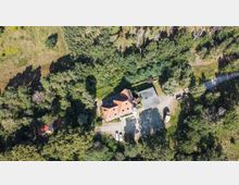Luftaufnahme eines Hauses mit roten Dächern und einem Nebengebäude, umgeben von dichtem Wald und Wiesen. Vor dem Haus befindet sich ein Kiesweg mit geparkten Autos.
