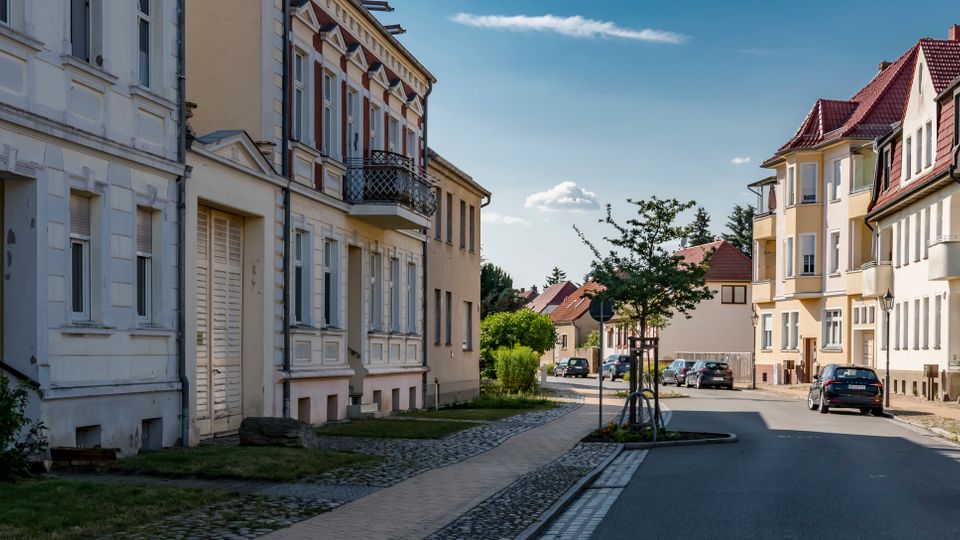 Wohnstraße in einem Wohnviertel mit mehrgeschossigen Altbauten und modernen Wohnhäusern, teils mit roten Ziegeldächern. Eine asphaltierte Straße mit Gehweg, Kopfsteinpflasterrand, jungen Straßenbäumen und parkenden Autos führt in eine leichte Kurve.