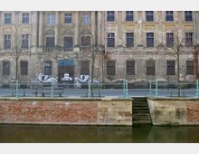 Fassade eines alten, verfallenen Gebäudes mit hohen Fenstern und Säulen, teilweise mit Graffiti bedeckt. Im Vordergrund eine Wasserfront mit gemauerter Uferbefestigung sowie eine Promenade mit grüner Metallgeländerung, Bänken und kahlen Bäumen.