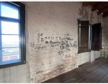 Eine Innenaufnahme eines alten Gebäudes mit weißen, teils abblätternden Ziegelwänden, auf denen handschriftliche Notizen oder Zeichnungen sichtbar sind. Links ist ein Fenster mit dunklem Metallrahmen, durch das Tageslicht einfällt, während rechts hölzerne Fensterläden geöffnet sind. Der Boden besteht aus dunklem Holz.