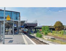 S-Bahnhof mit überdachtem Bahnsteig, auf dem Fahrgäste warten, Gleise und ein einfahrender Zug. Im Hintergrund eine Brücke mit Straßenbahnen und der Berliner Fernsehturm in der Ferne sichtbar.
