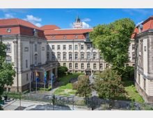 Historisches Gebäude mit hellem Mauerwerk und rotem Ziegeldach, umgeben von einem begrünten Innenhof mit Bäumen und blühenden Sträuchern. Im Vordergrund stehen fünf Fahnenmasten vor einem Eingangsbereich mit Säulen.