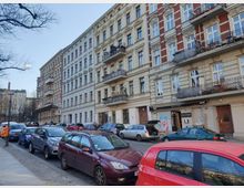 Straßenszene mit Altbau-Wohnhäusern im Berliner Stil, gekennzeichnet durch verputzte und rote Ziegelfassaden mit großen Fenstern und Balkonen mit schmiedeeisernen Geländern. Am Straßenrand stehen geparkte Autos, und im Hintergrund sind Bäume sowie ein weiteres Gebäude sichtbar.