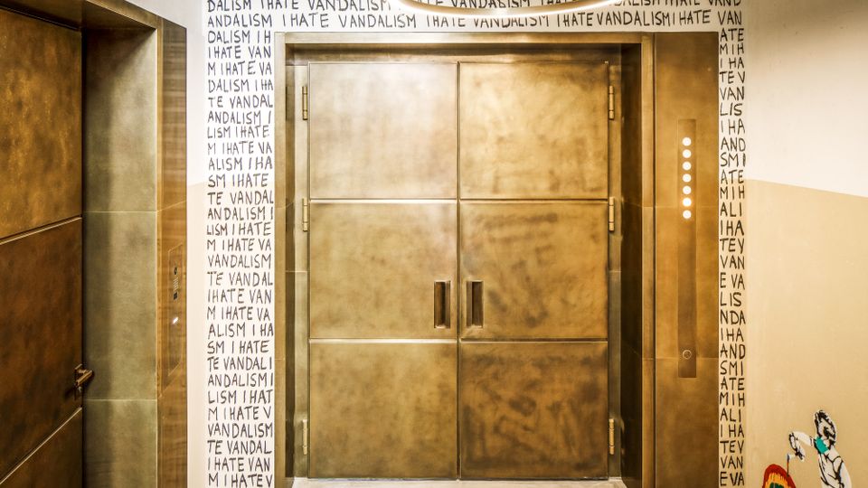 Flur mit großen, goldfarbenen Aufzugstüren und einem ringförmigen Deckenlicht. Die Wände sind mit wiederholtem Schriftzug „I HATE VANDALISM“ beschriftet, daneben befinden sich Bedientasten und eine kleine Wandzeichnung.