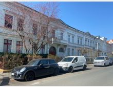 Straßenansicht mit einer Reihe historischer, weiß gestrichener Gebäude mit großen Fenstern und roten Akzenten, flankiert von blattlosen Bäumen. Vor den Gebäuden parken mehrere Autos, darunter ein schwarzer Kleinwagen und zwei weiße Transporter.