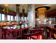 Ein stilvoll eingerichteter Innenraum eines asiatischen Restaurants mit roten Wänden, glänzenden Marmorsäulen und einem großen Kronleuchter. Die Einrichtung umfasst Holzmöbel mit roten Polstern, traditionelle Keramiken und grüne Lampenschirme als Dekoration.