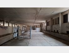 Innenansicht einer großen, verlassenen Industriehalle mit niedriger Betondecke, Stützpfeilern und seitlichen Trennwänden mit Graffiti. Der lange Gang führt zu einem vergitterten Bereich am Ende; auf dem Boden liegen verstreute Abfälle und einzelne Platten.