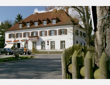 Zweistöckiges weißes Gebäude mit rotem Satteldach, mehreren Dachgauben und Holzfensterläden, im Erdgeschoss mit einer Bäckerei. Im Vordergrund eine Straße, parkende Autos und ein Holzzaun; im Hintergrund Bäume.