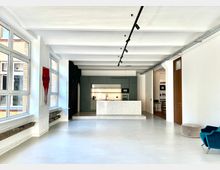 Moderner Innenraum mit offener Küche, die eine graue Einbauschrankwand und eine zentrale Kücheninsel mit weißer Marmoroptik enthält. Große Fenster lassen viel Tageslicht herein, helle Wände und ein polierter Betonboden prägen den Raum, ergänzt durch einen türkisfarbenen Sessel und einen beigefarbenen Hocker.