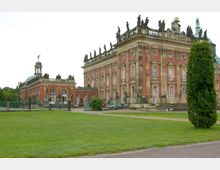 Barockes Schloss mit roter Fassade, Sandsteinsäulen und Zierelementen, flankiert von Statuen und einem Kuppelbau mit goldener Figur an der Spitze. Im Vordergrund eine Rasenfläche und ein kleiner Weg.