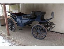 Eine historische, blaue Pferdekutsche mit großen Holzrädern und kunstvollen Details, abgestellt auf einem gepflasterten Untergrund unter einem Überdach, neben einer hellen Wand.