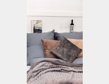 Ein gemütlich hergerichtetes Bett mit mehreren Kissen in unterschiedlichen Farben und Mustern, darunter Grau-, Braun- und Metallic-Töne. Im Hintergrund auf einer weißen Wand befinden sich zwei Weingläser, eine Weinflasche und ein aufgestelltes Buch mit Berglandschaftsmotiv.