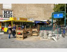 Eine Straßenszene am U-Bahnhof Rosenthaler Platz in Berlin mit einem gelben Imbissstand namens "Rosenback" und einer Baustelle im Vordergrund, abgesichert durch Absperrbänder und Holzbarrieren. Im Hintergrund sind eine backsteinerne Wand, weitere Stände und Fahrradständer zu sehen.