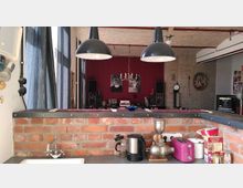 Küche mit einer Arbeitsplatte aus grauem Stein und einer Backsteinmauer im Vordergrund. Im Hintergrund ein Wohnraum mit roten Wänden, zwei großen Lautsprechern und einer Musikanlage; darüber hängt ein Kronleuchter, und an den Wänden sind Poster sowie eine Uhr und religiöse Kunstwerke zu sehen.