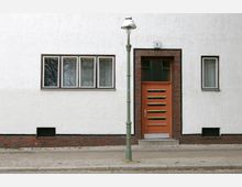 Fassade eines Wohnhauses mit weiß verputzter Wand, einer orangefarbenen Eingangstür mit horizontalen Glaseinsätzen, flankiert von roten Klinkersteinen, sowie mehreren Fenstern. Vor dem Gebäude steht eine grüne Straßenlaterne.