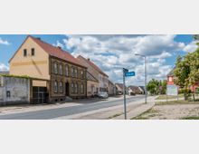Straßenszene in einem Dorf mit Wohnhäusern entlang einer asphaltierten Straße. Zu sehen sind ein historisches Backsteinhaus, Gebäude mit roten Dächern und eine Straßenschild mit der Aufschrift „Dorfstraße“, daneben Bäume und eine Infotafel.