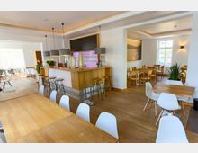 Modernes Café oder Restaurant mit hellem Holzfußboden, minimalistischen Holztischen und weißen Stühlen. Im Zentrum befindet sich eine Theke mit Holzfront, darüber hängen drei graue Lampen; im Hinterraum sind weitere Sitzbereiche sichtbar.