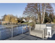 Terrasse mit Geländer und Sitzmöbeln aus Rattan, angrenzend an einen ruhigen Fluss mit Schilf und kleinen Anlegestegen. Im Hintergrund sind Bäume, Wohnhäuser und ein klarer blauer Himmel sichtbar.