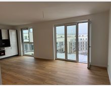 Wohnzimmer einer modernen Wohnung mit hellen Wänden und Holzfußboden. Große bodentiefe Fenster mit Blick auf einen Balkon und umliegende Wohngebäude; in der linken Ecke befindet sich eine weiße Einbauküche mit schwarzen Akzenten.
