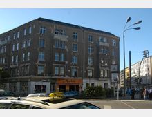 Straßenecke mit einem vierstöckigen Wohn- und Geschäftshaus in grauem Putz, teilweise reparaturbedürftig; im Erdgeschoss befinden sich mehrere Läden, darunter ein Café mit orangefarbener Markise. Im Vordergrund parkt ein Taxi, und im Hintergrund sind Fußgänger sowie weitere mehrstöckige Gebäude sichtbar.