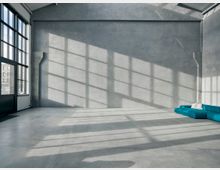 Industrieller Innenraum mit grauen Betonwänden, großen Fenstern, die rechteckige Licht- und Schattenmuster auf dem Boden erzeugen, und einer türkisfarbenen Eckcouch mit weißen Kissen in einer Ecke.