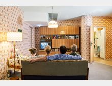 Ein Wohnzimmer im Stil der 1970er Jahre mit gemusterten Tapeten, einer Holz-Schrankwand, einem Röhrenfernseher und einer Stehlampe. Im Vordergrund sitzt eine vierköpfige Familie auf einem Sofa und schaut fern; im Hintergrund führt eine geöffnete Tür in ein Badezimmer.